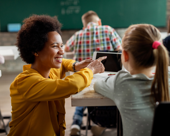 Smiling teacher kneeling beside a student’s desk, pointing at a tablet and offering guidance in a bright classroom with a chalkboard in the background
