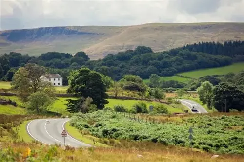 Curving country road through lush Welsh hills and farmland, with a white farmhouse and distant Brecon Beacons under a bright, cloudy sky.