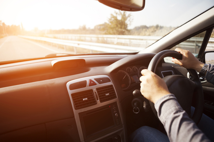 Driver’s hands on a right‑hand‑drive steering wheel on a sunny open road, car interior view