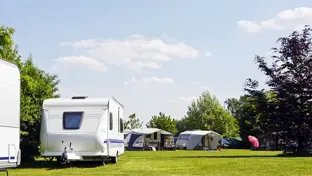 Sunny campsite with caravans and awnings on a wide green lawn under a blue sky with scattered clouds.