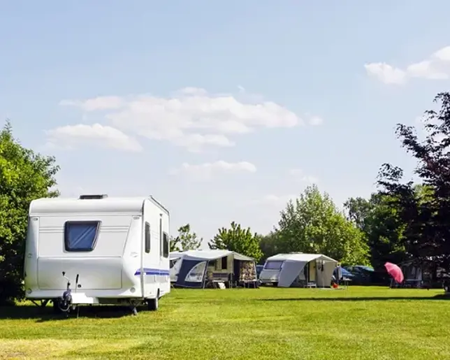 Sunny campsite with caravans and awnings on a wide green lawn under a blue sky with scattered clouds.