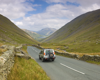 A silver SUV drives along the winding Kirkstone Pass, surrounded by dry stone walls and lush fells under a partly cloudy sky.