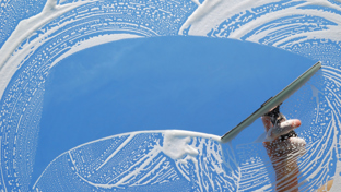 Hand using a squeegee to clear soapy suds from a window, revealing a bright blue sky