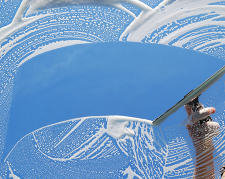 Hand using a squeegee to clear soapy suds from a window, revealing a bright blue sky