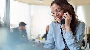 A smiling woman in a blue blouse talks on the phone at her office desk