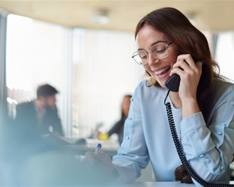 A smiling woman in a blue blouse talks on the phone at her office desk