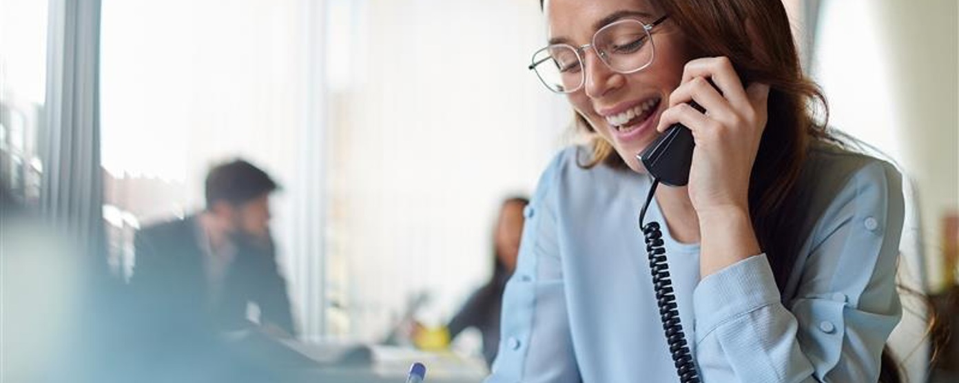 A smiling woman in a blue blouse talks on the phone at her office desk