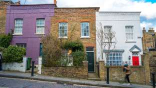 Three terraced houses in purple, brick, and white with small front gardens on a cobblestone street.