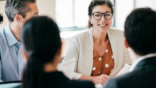 A smiling female professional in glasses leads a meeting, engaging her colleagues in a collaborative discussion.