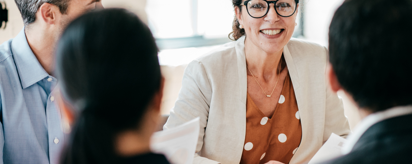A smiling female professional in glasses leads a meeting, engaging her colleagues in a collaborative discussion.