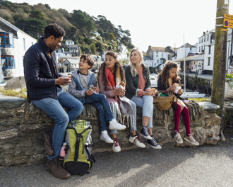 Mixed family sitting on a harbour wall in a coastal village, sharing lunch and chatting together