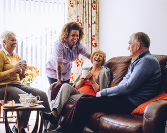 A group of elderly care home residents sit together with a caregiver, laughing and chatting over tea in a bright, cheerful living room