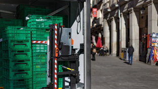 The back of a delivery truck is open, revealing stacked green plastic crates on wheeled platforms inside.