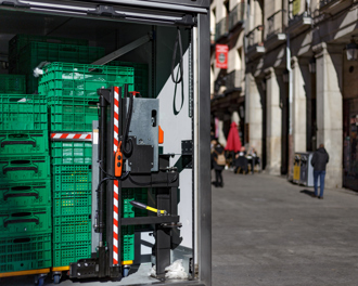The back of a delivery truck is open, revealing stacked green plastic crates on wheeled platforms inside.