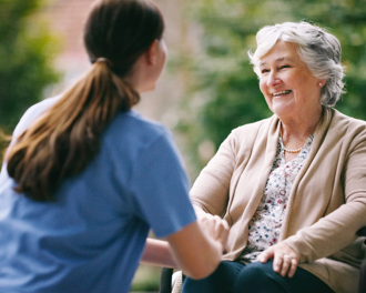 Smiling elderly woman in a wheelchair talking with a caregiver outdoors.