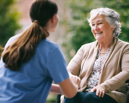 Smiling elderly woman in a wheelchair talking with a caregiver outdoors.