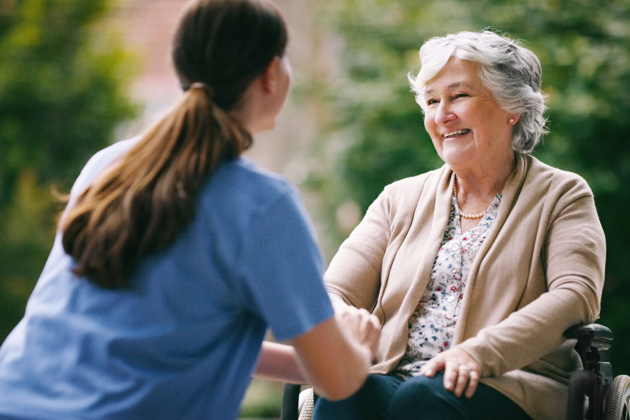 Smiling elderly woman in a wheelchair talking with a caregiver outdoors.