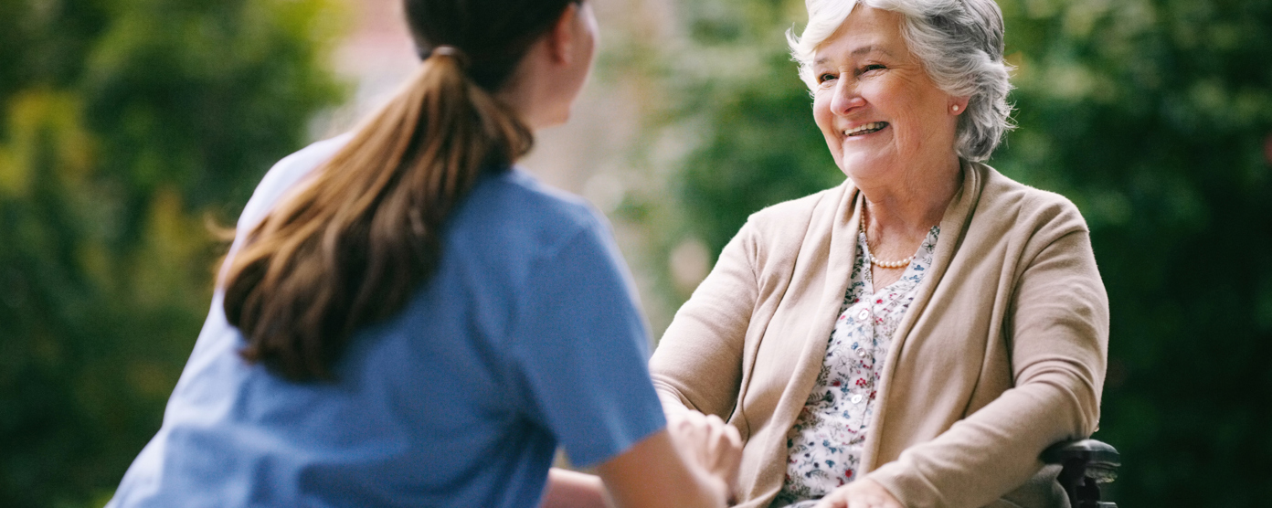 Smiling elderly woman in a wheelchair talking with a caregiver outdoors.