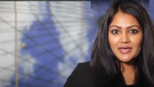 A woman with long, dark hair and medium skin tone is speaking or presenting indoors.