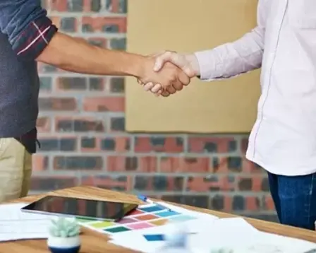 Two people standing and shaking hands across a desk, suggesting a professional agreement or business deal.