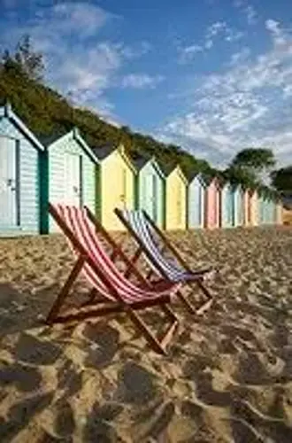 A row of colorful beach huts lines the sandy shore, with two striped deck chairs in the foreground, facing the bright blue sky scattered with fluffy clouds.