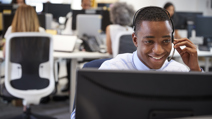 A smiling young man wearing a headset is sitting at a desk in a busy office environment, looking at a computer monitor.