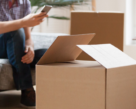 A person is seated on a grey couch in a well-lit living room surrounded by several cardboard boxes.
