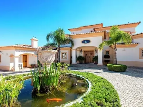 A grand villa with a white and terracotta exterior, featuring a circular driveway leading to the main entrance.