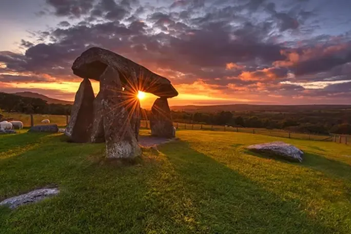 Ancient stone dolmen structure on a grassy field at sunset, with sun rays shining through the stones and dramatic clouds in the sky.