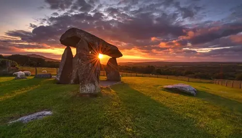 Ancient stone dolmen structure on a grassy field at sunset, with sun rays shining through the stones and dramatic clouds in the sky.