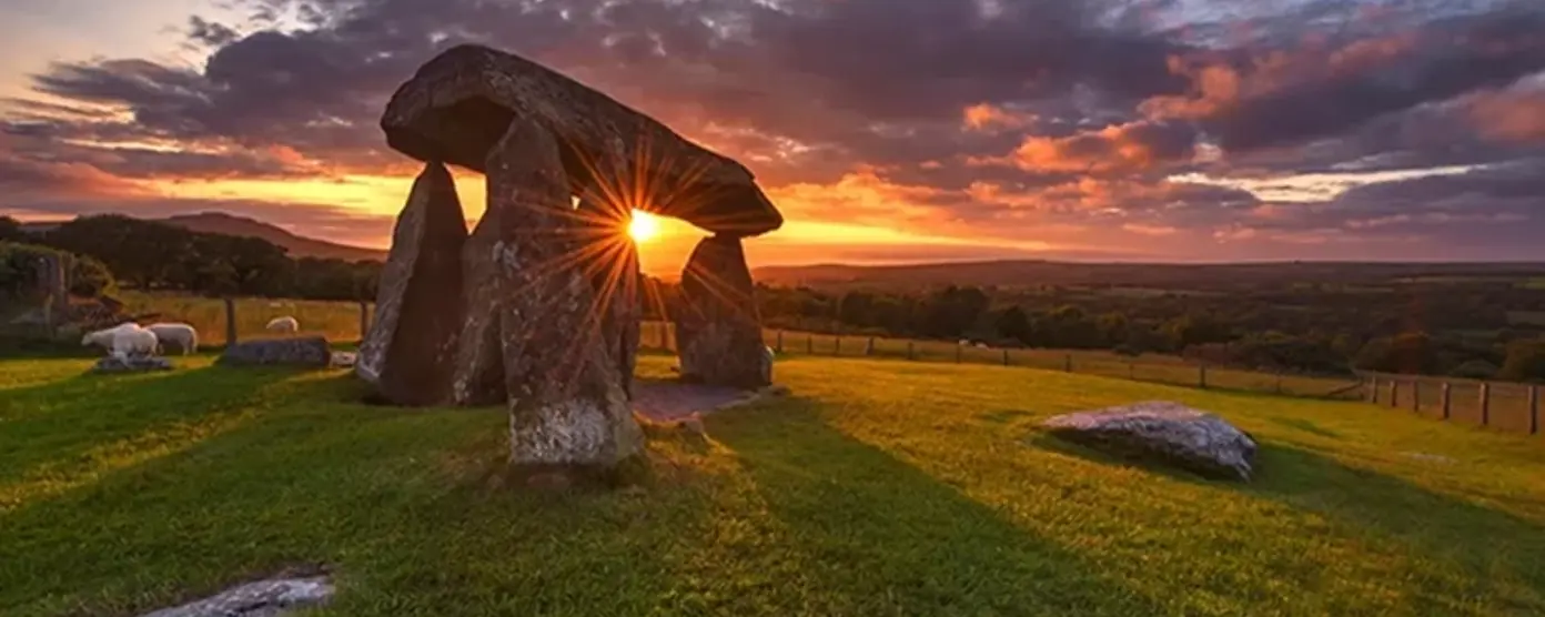 Ancient stone dolmen structure on a grassy field at sunset, with sun rays shining through the stones and dramatic clouds in the sky.