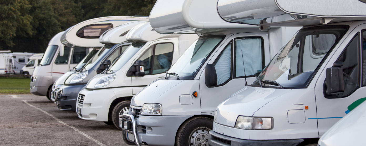 Row of motorhomes parked side by side at a campsite, angled front view with trees in background