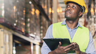 A male worker wearing a yellow hard hat and a high-visibility vest is inspecting a warehouse and also holding a clipboard and looking upward with a thoughtful expression.