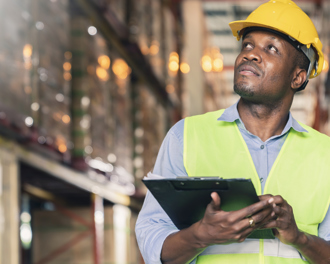 A male worker wearing a yellow hard hat and a high-visibility vest is inspecting a warehouse and also holding a clipboard and looking upward with a thoughtful expression.