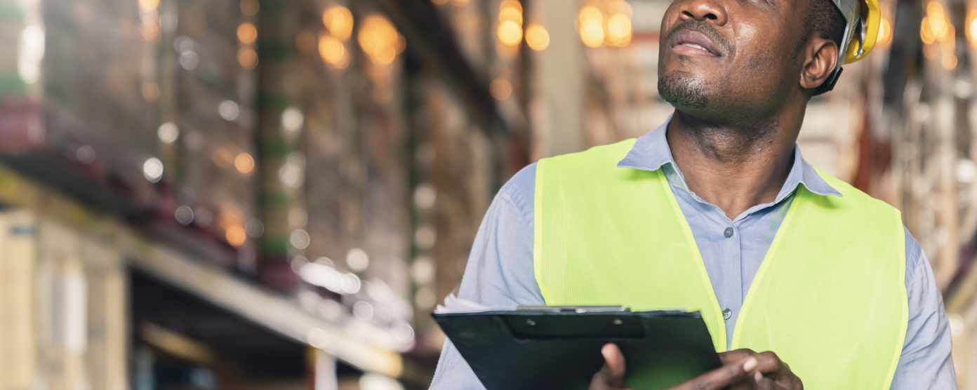 A male worker wearing a yellow hard hat and a high-visibility vest is inspecting a warehouse and also holding a clipboard and looking upward with a thoughtful expression.