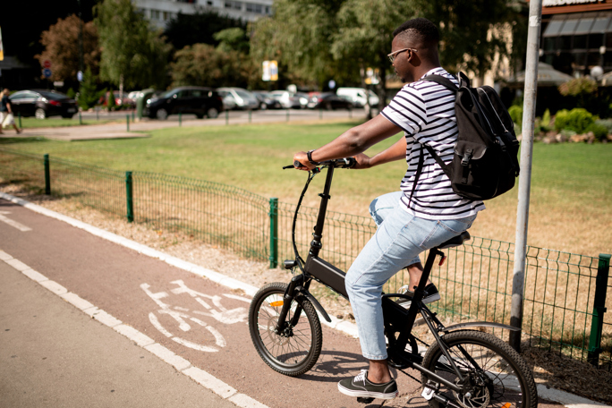 A man rides a black electric bike on a dedicated bike path marked with a white bicycle symbol. He wears a white and black striped T-shirt, light blue jeans, and black sneakers.