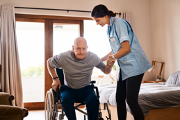 A female caregiver in a light blue uniform assists an elderly man as he rises from a wheelchair in a well-lit bedroom.