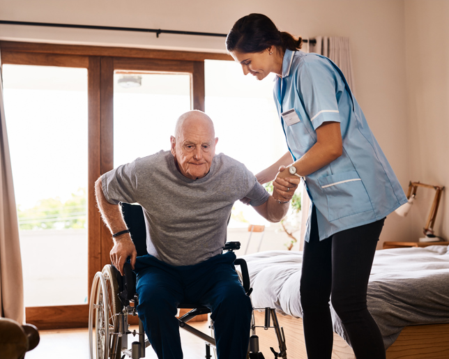 A female caregiver in a light blue uniform assists an elderly man as he rises from a wheelchair in a well-lit bedroom.