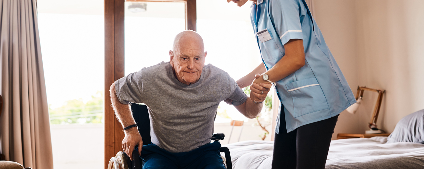 A female caregiver in a light blue uniform assists an elderly man as he rises from a wheelchair in a well-lit bedroom.