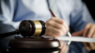 A close-up image of a wooden judge's gavel with a golden band resting on a sound block on a reflective surface.