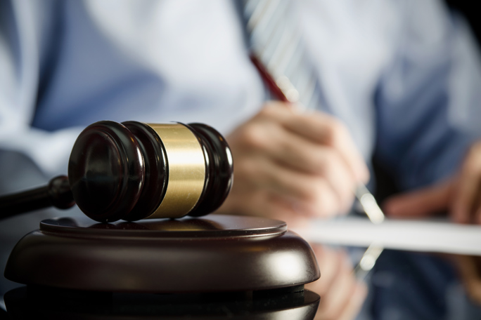 A close-up image of a wooden judge's gavel with a golden band resting on a sound block on a reflective surface.