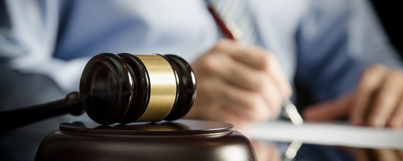 A close-up image of a wooden judge's gavel with a golden band resting on a sound block on a reflective surface.