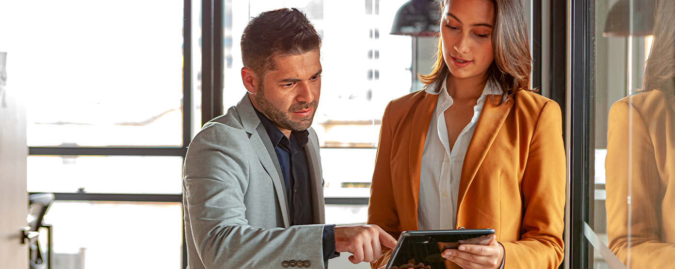 Two colleagues smartly dressed are looking at a tablet together, the male pointing at the device, in an office environment.