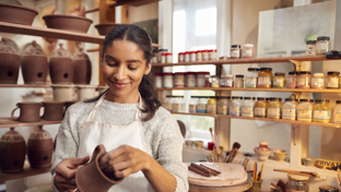 A woman wearing a white apron and a light sweater is smiling while shaping a clay pot in a pottery studio.