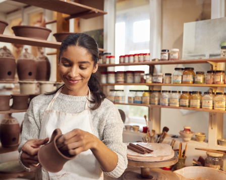 A woman wearing a white apron and a light sweater is smiling while shaping a clay pot in a pottery studio.