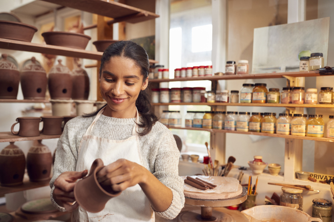 A woman wearing a white apron and a light sweater is smiling while shaping a clay pot in a pottery studio.