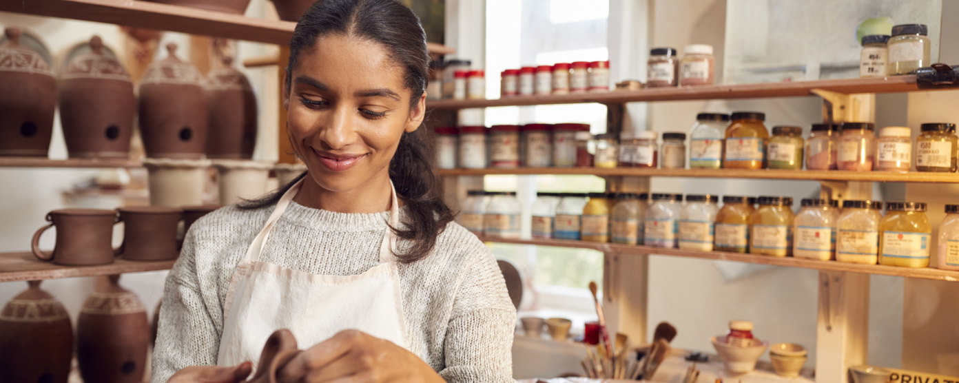 A woman wearing a white apron and a light sweater is smiling while shaping a clay pot in a pottery studio.