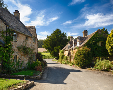 Village street with traditional stone cottages and green foliage under a blue sky.