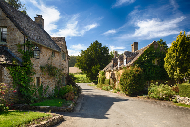 Village street with traditional stone cottages and green foliage under a blue sky.