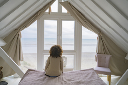 A person sits on a bed in a cozy attic bedroom with large triangular windows, looking out at the beach and ocean.
